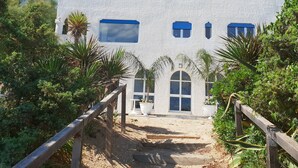 Un bâtiment blanc aux fenêtres encadrées de bleu, un escalier en bois menant à l’entrée et une végétation luxuriante tout autour.