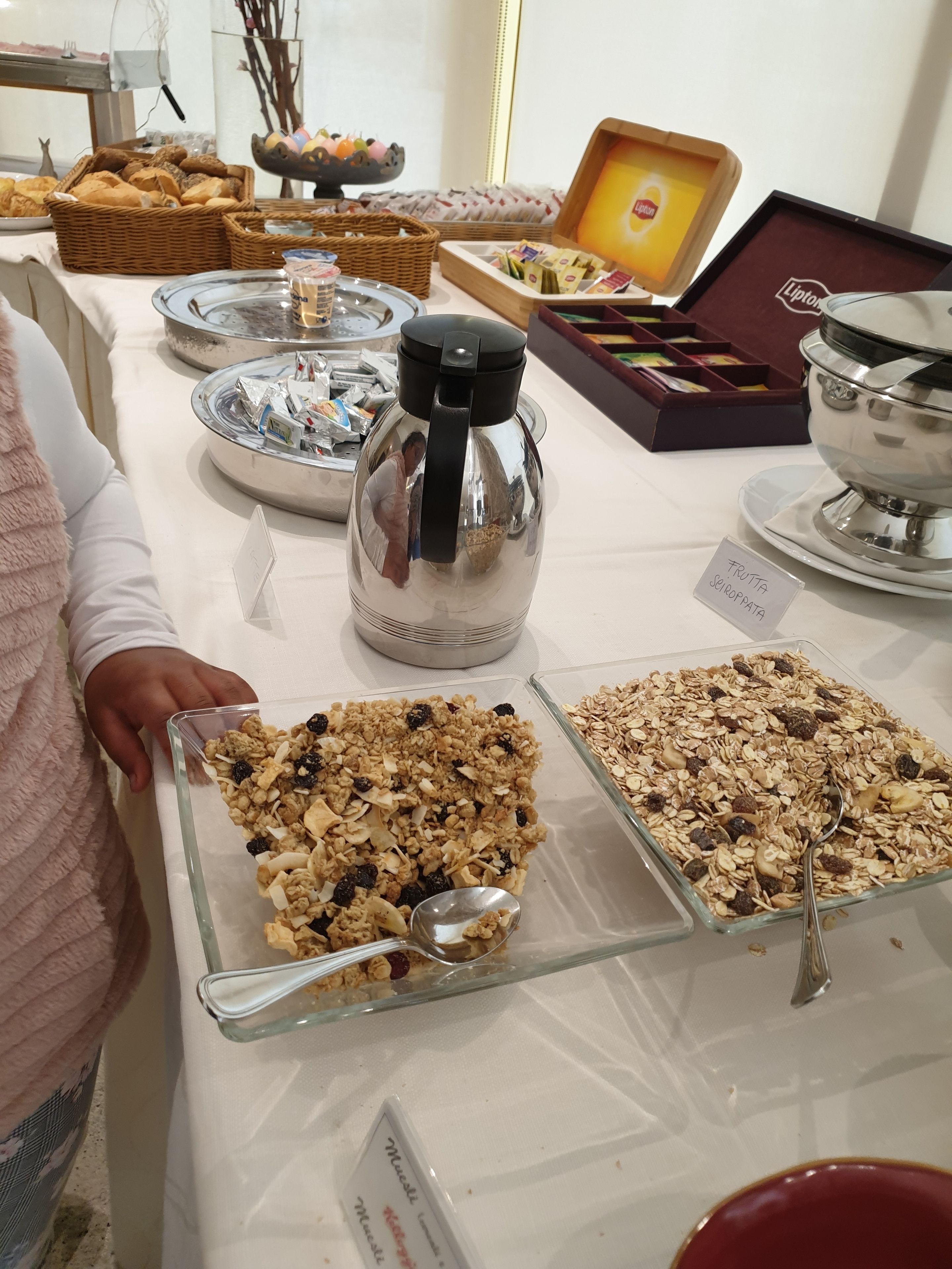 A buffet table with various food items, including a glass container labeled "FRUTTA SIOROPPA," a metal coffee pot, and a bowl of granola.