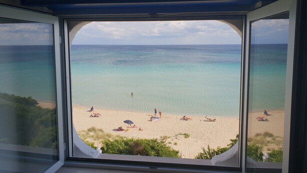 Une vue sur la plage, à travers une fenêtre, avec une eau turquoise et limpide et une plage de sable.