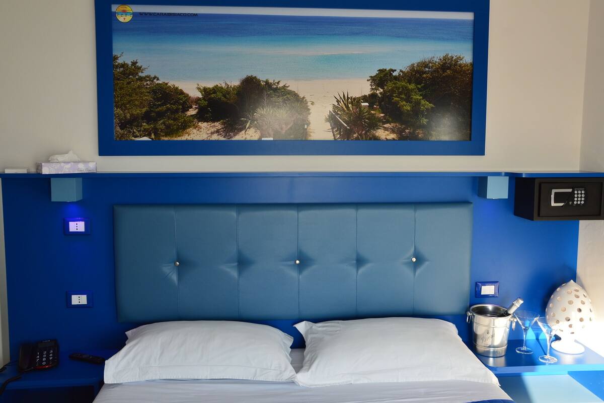 A hotel room with a large framed beach view, a blue headboard, a bed with white linens, a bedside table with a bucket and glasses, and a phone on the nightstand.