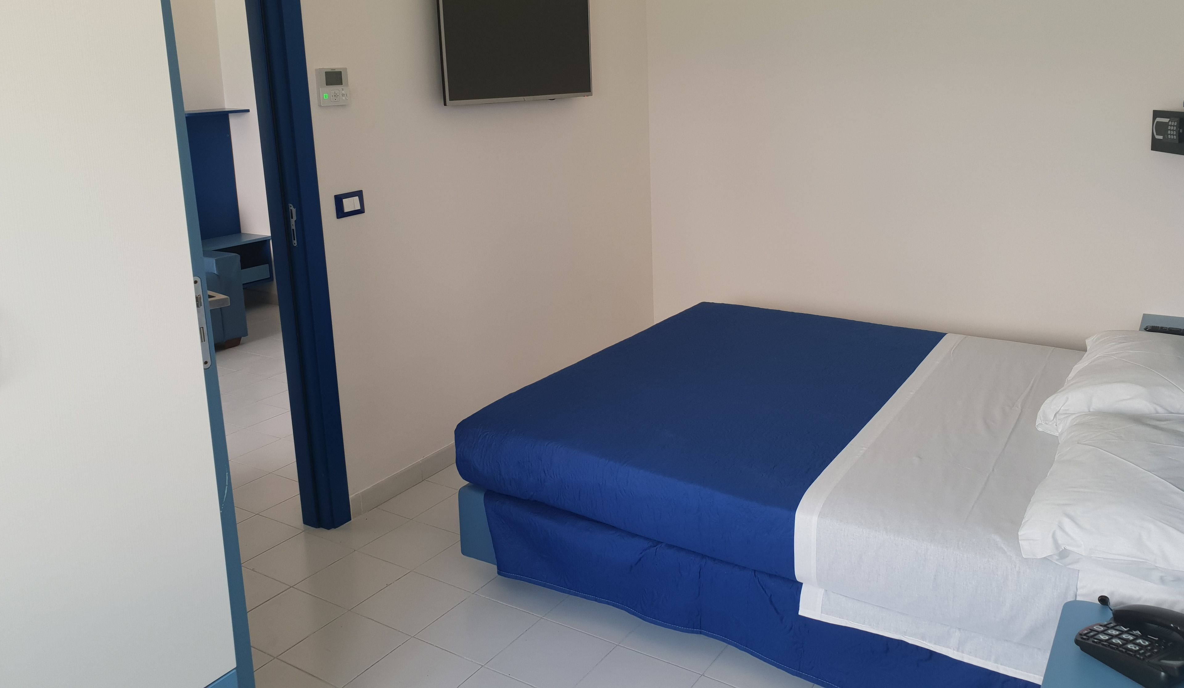 Ein Zimmer mit einem Bett, einem Schreibtisch und einem Stuhl. Eine Tür führt in einen weiteren Raum.