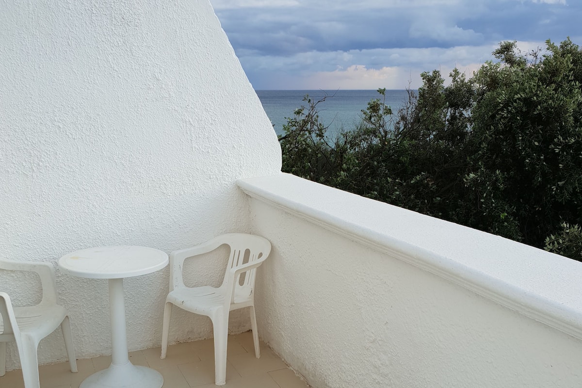 A balcony with white plastic furniture, including a round table and two chairs, overlooking a body of water and greenery.