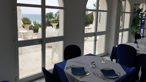 Un espace repas avec des tables préparées pour deux personnes, de grandes fenêtres en arc de cercle et une vue sur la mer, ainsi que des sièges à l'extérieur.