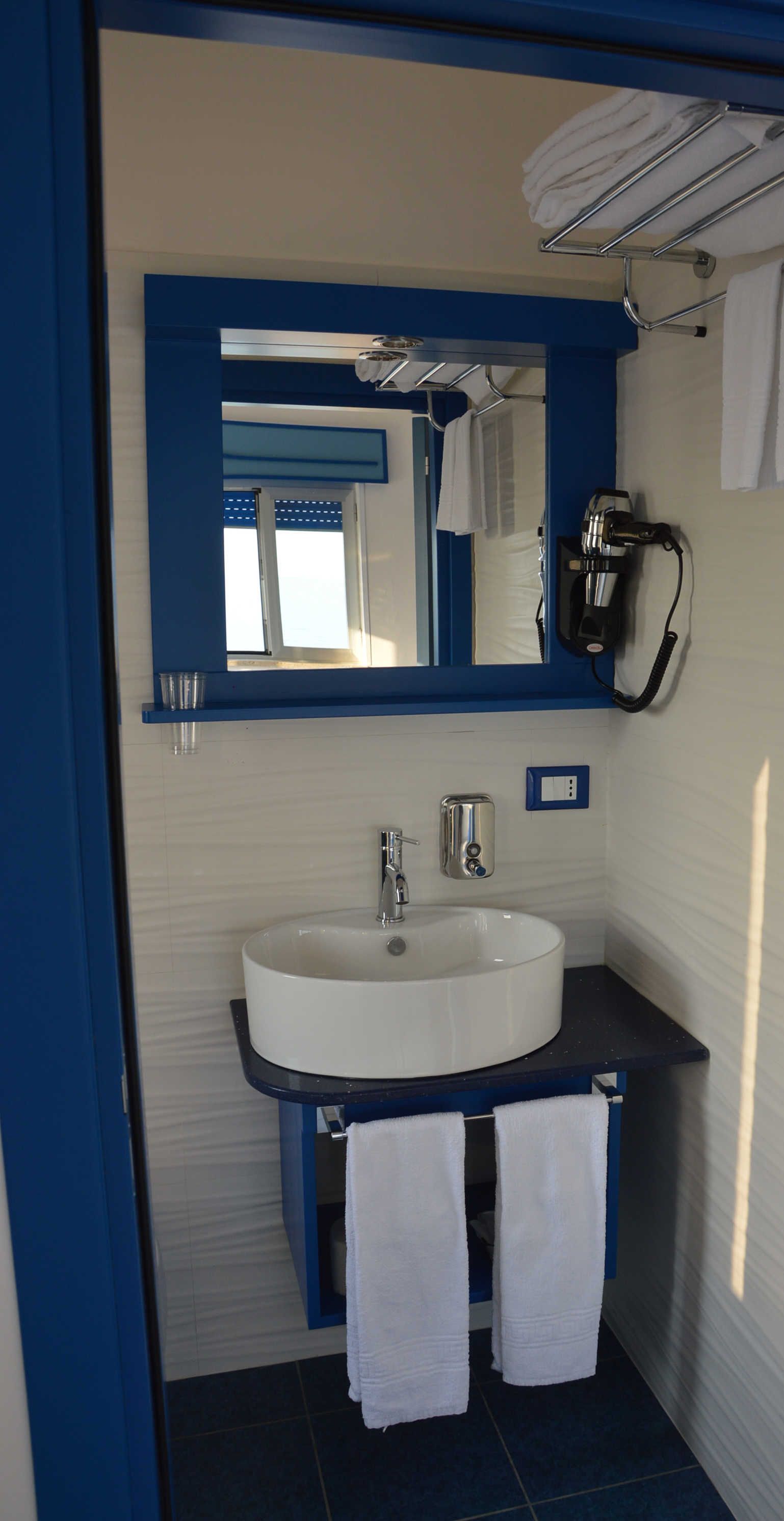 A bathroom with a white sink, blue countertop, and a mirror above it. There is a towel rack with towels and a hairdryer on the wall.