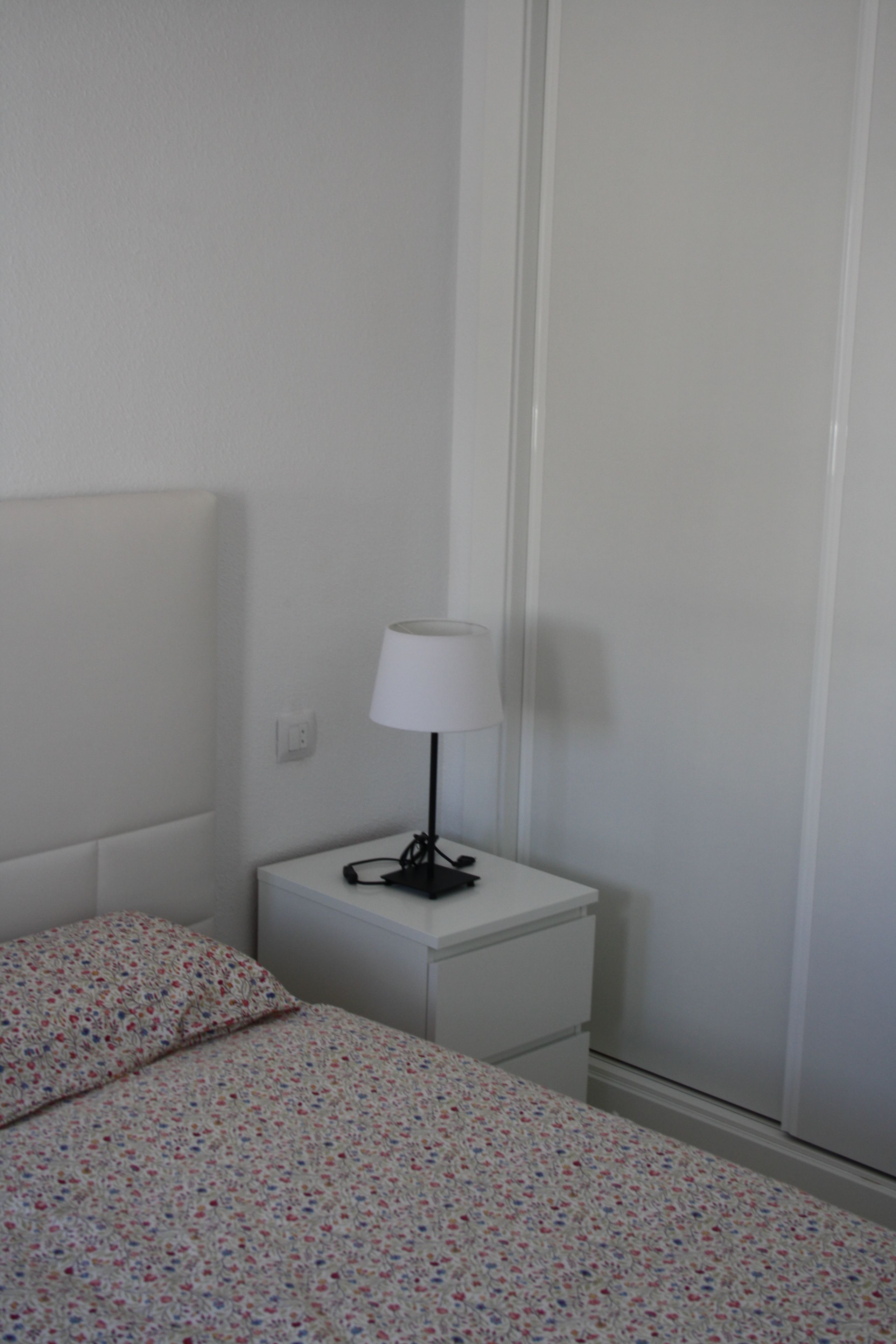 Un dormitorio con una cama, una mesita de noche con lámpara y una pared blanca.