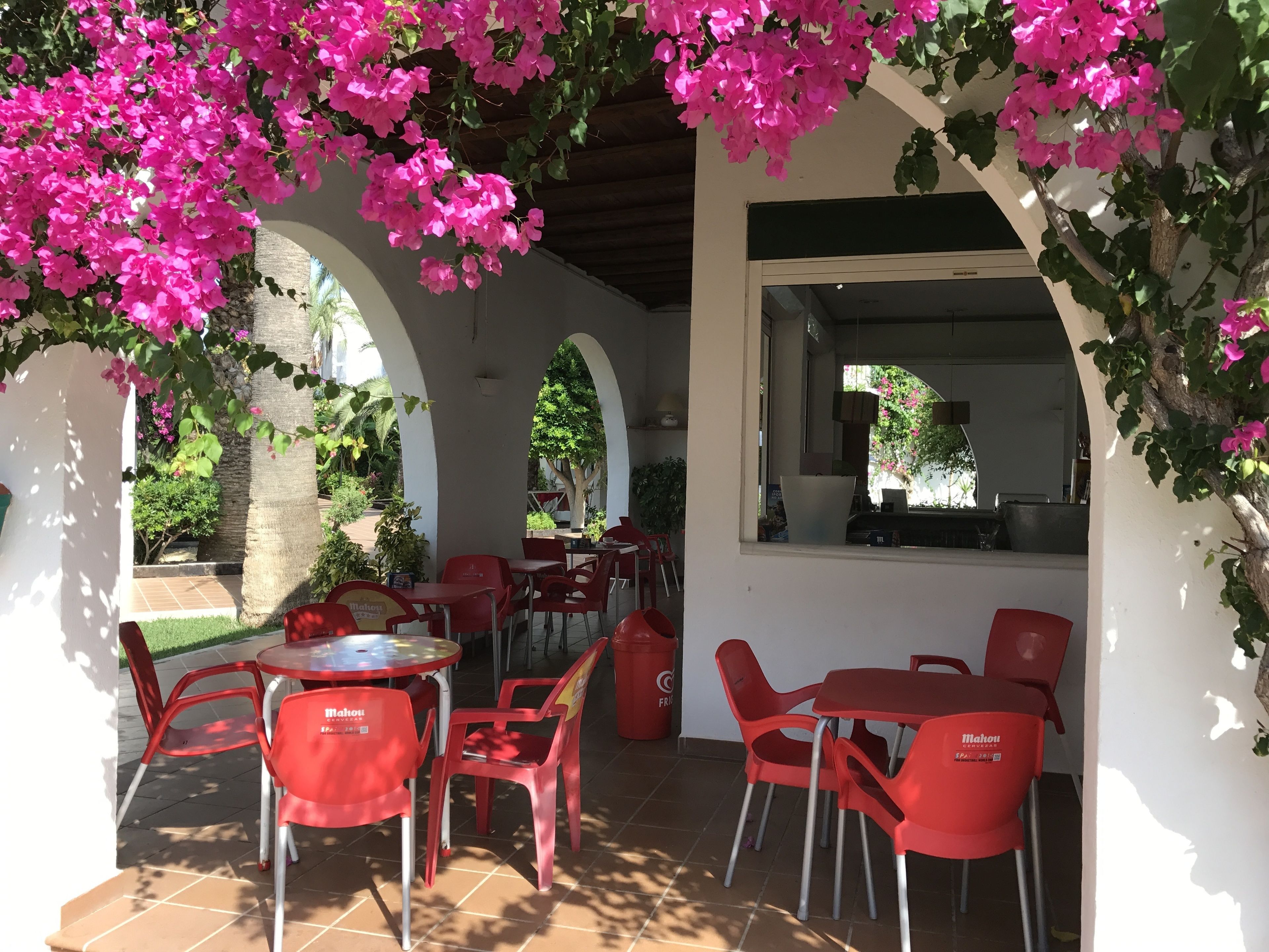 Un área de café al aire libre con sillas y mesas de plástico rojo, rodeada de arcos y flores de buganvilla rosa.