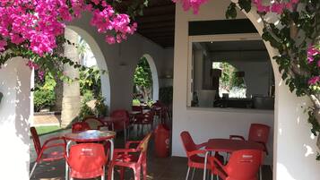 Un área de café al aire libre con sillas y mesas de plástico rojo, rodeada de arcos y flores de buganvilla rosa.