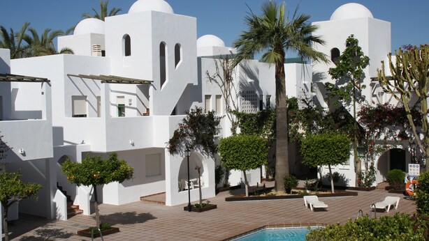 A pool area with white buildings, palm trees, and lounge chairs. - Apartamentos Torrelaguna (Vera)