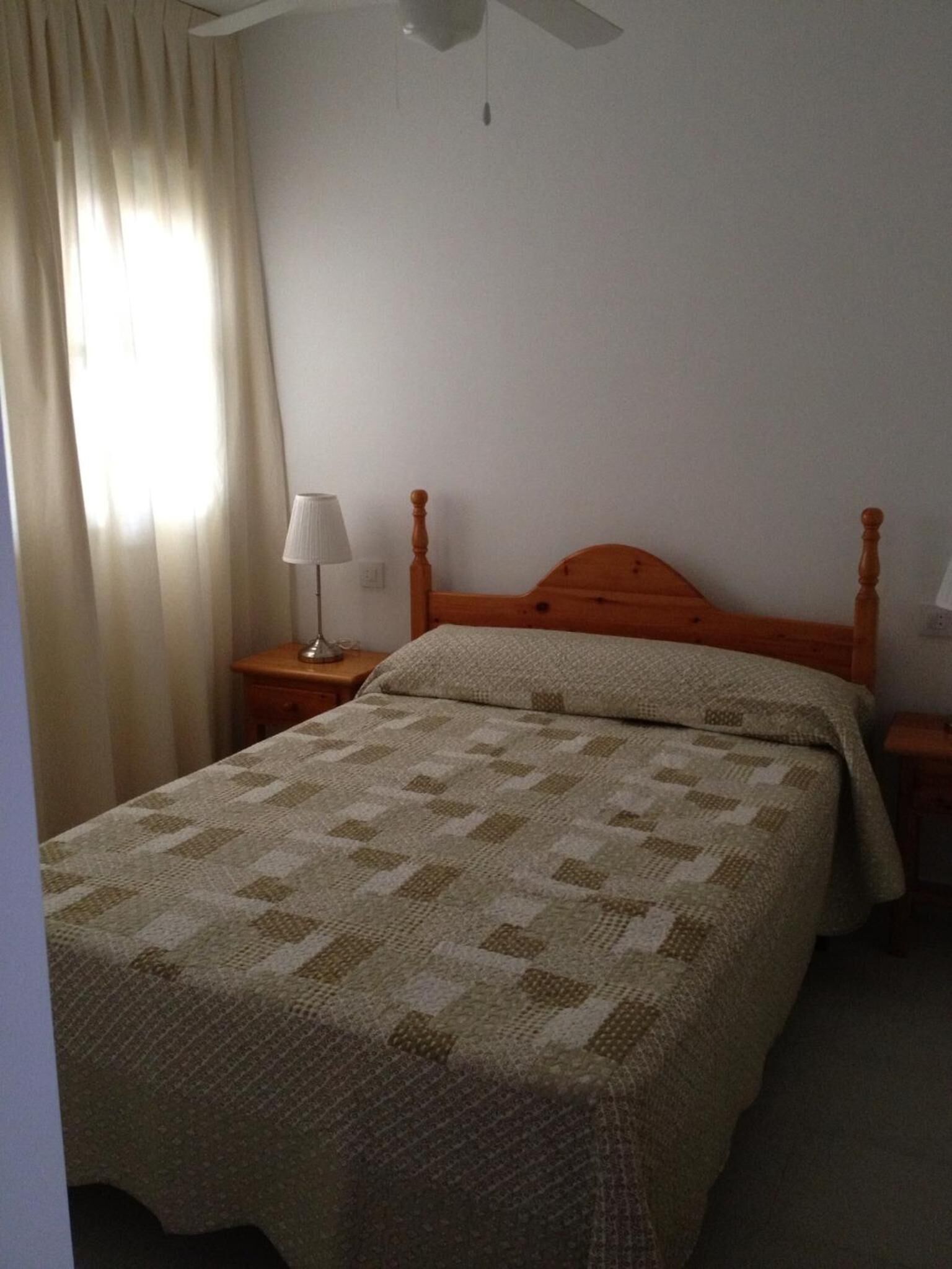 Una cama individual con cabecera de madera, una mesita de noche con lámpara y una ventana con cortinas.
