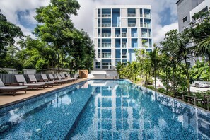 A swimming pool with lounge chairs, surrounded by trees and a modern building with balconies in the background. - DeeProm Pattaya Hotel (Pattaya)