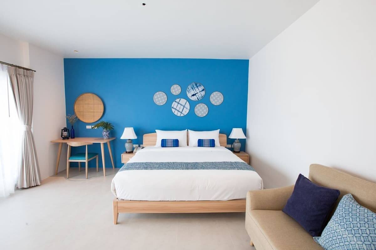 A modern bedroom with a blue accent wall, a wooden bed, a beige sofa, and a round mirror.