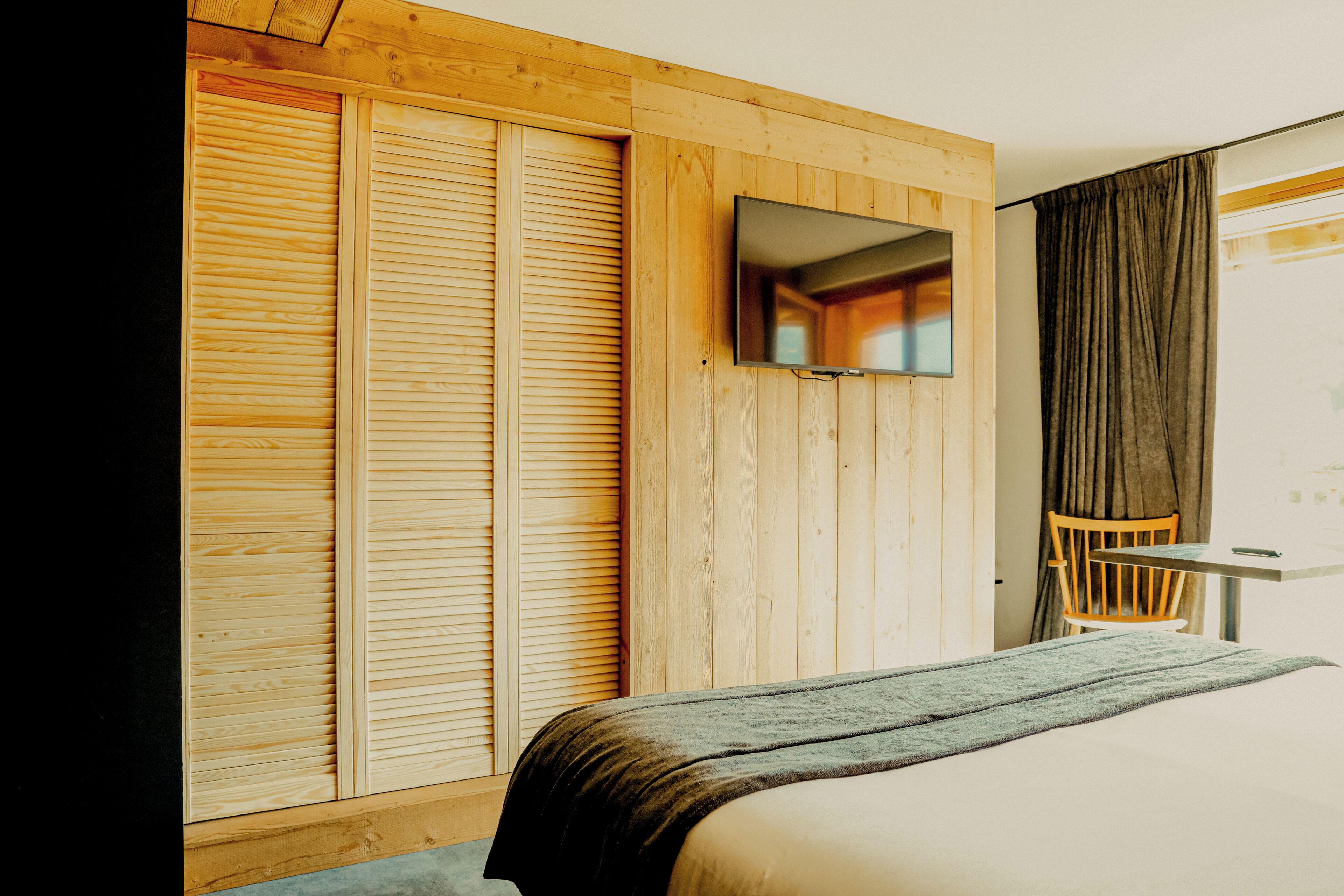 A hotel room with a bed, a wooden wardrobe, a television mounted on the wall, and a chair.