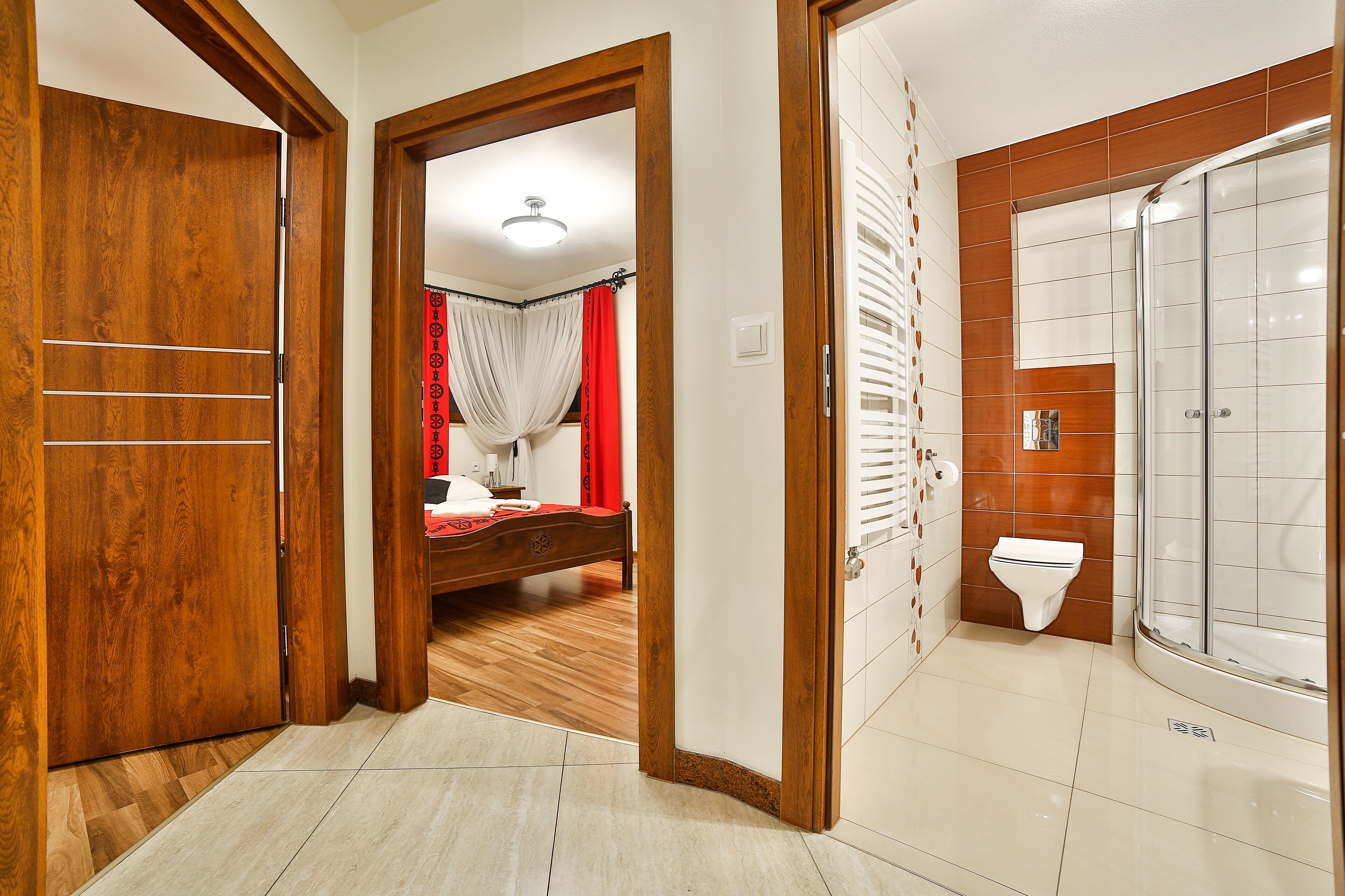 A hotel room with a wooden door, a bed with a red and white bedspread, a shower with a glass door, and a white toilet.