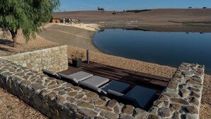 A stone-walled outdoor seating area with lounge chairs and a wooden deck overlooking a calm lake. - Monte Vale Organic Farm (Castro Verde)