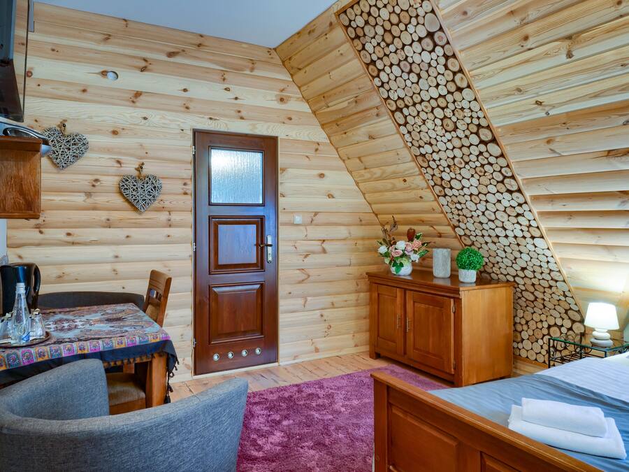 A cozy log cabin room with a wooden ceiling, a bed, a table with chairs, and a small cabinet.