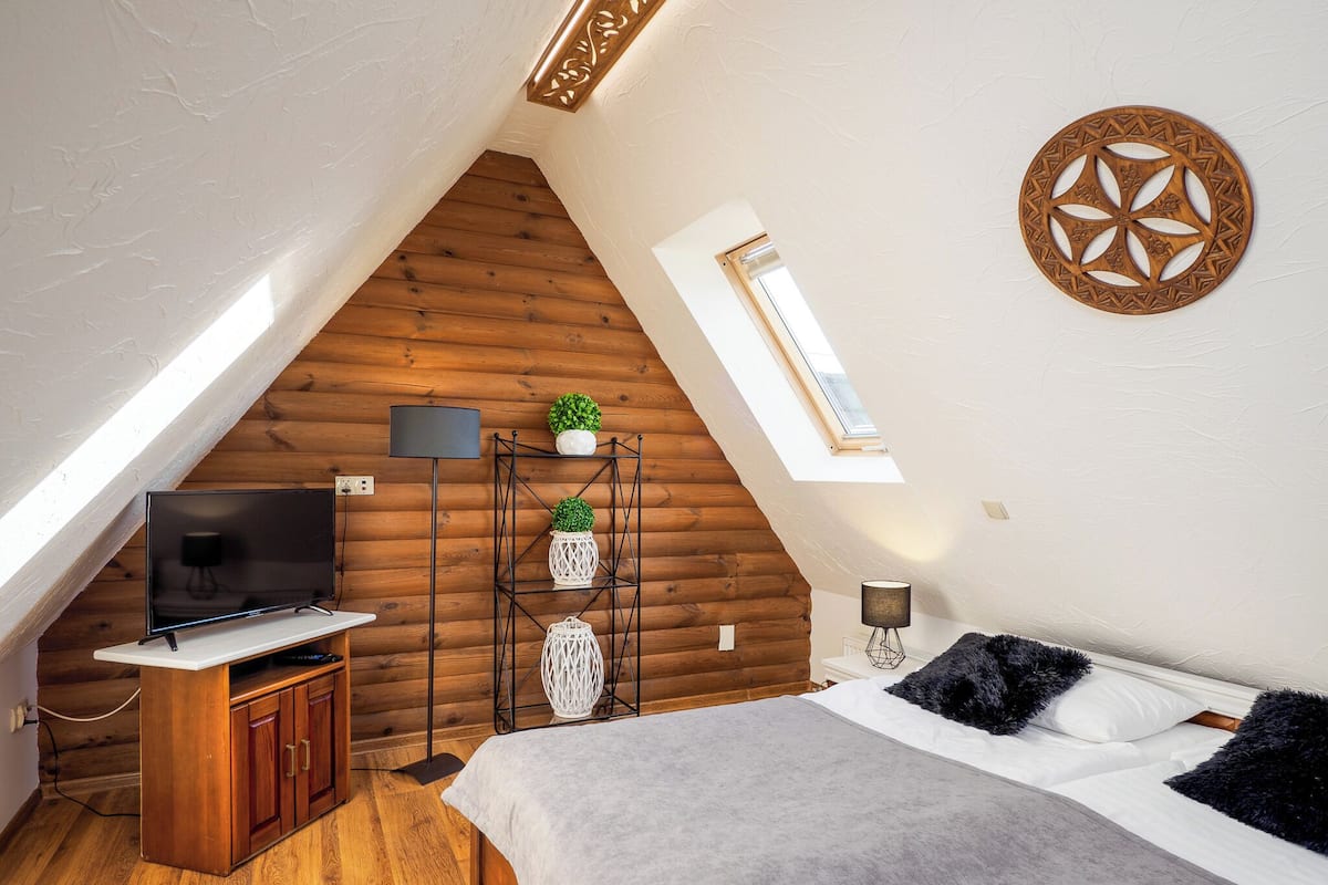 Une chambre confortable avec un plafond en pente, des murs en bois, un lit avec des oreillers de couleurs foncées et claires, un téléviseur posé sur un meuble en bois et une petite fenêtre.