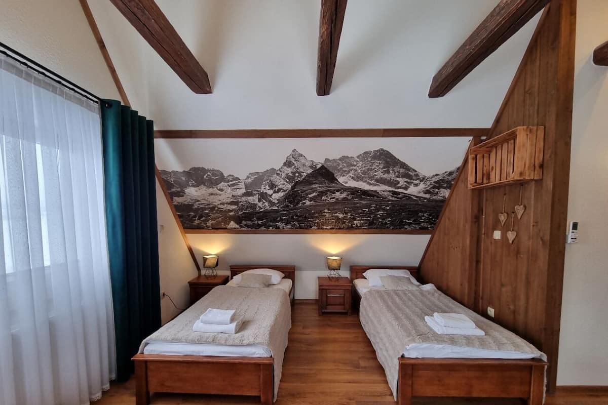 Une chambre avec deux lits en bois, une fresque représentant une montagne sur le mur, des poutres en bois au plafond et une fenêtre ornée de rideaux légers.