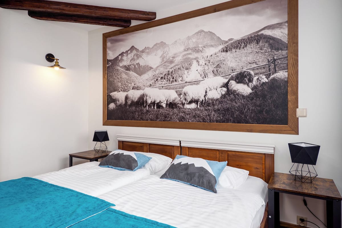Une chambre à coucher avec un grand tableau représentant un paysage de montagne, une tête de lit en bois, un lit avec une literie blanche et bleue, et deux tables de chevet avec des lampes.