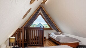 Een kleine, driehoekige slaapkamer op zolder met twee bedden, een houten ledikant en uitzicht op een groen landschap door een groot raam.
