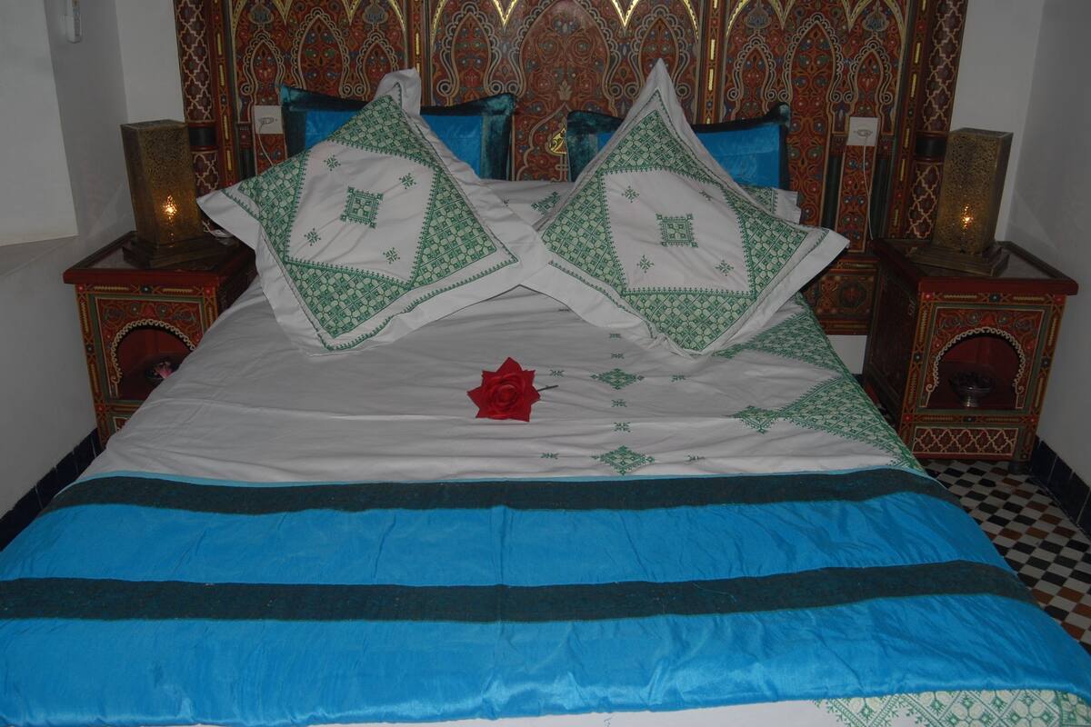 Una cama con una colcha azul y blanca, dos almohadas decorativas y una rosa roja.