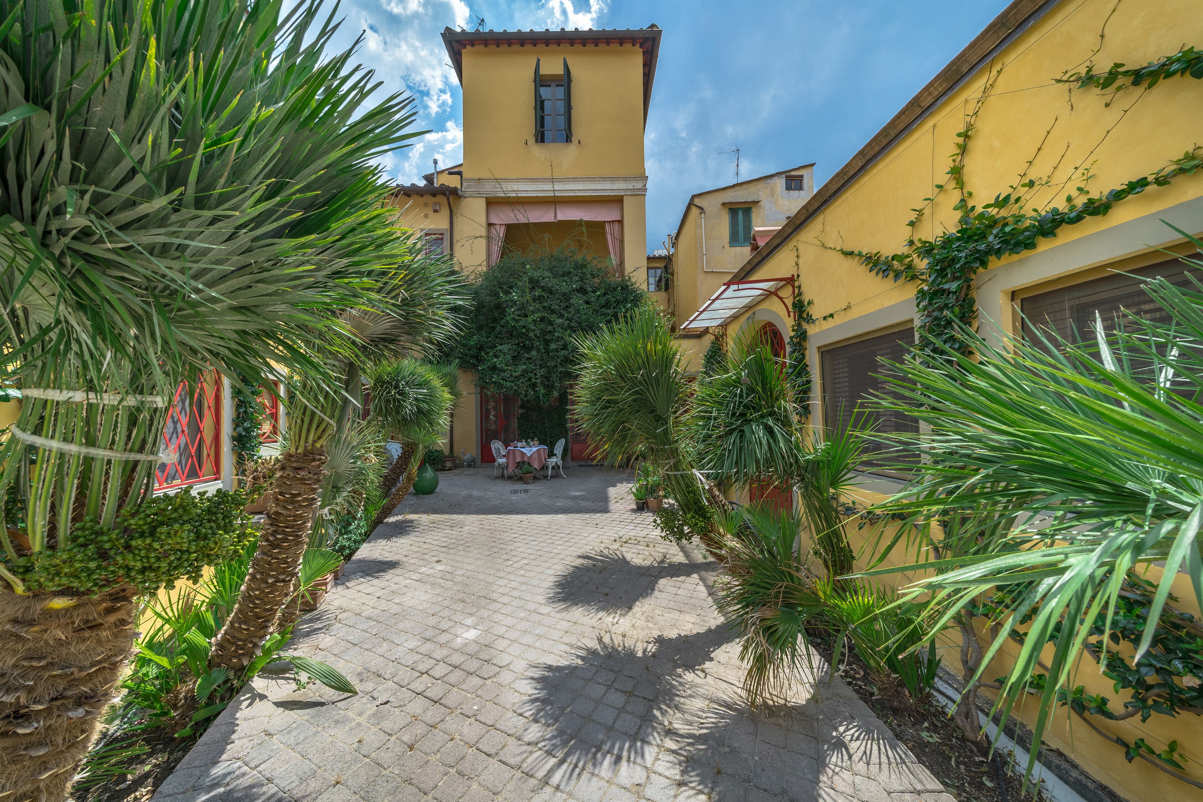 A courtyard with a paved walkway, surrounded by yellow buildings, palm trees, and a small table with chairs.