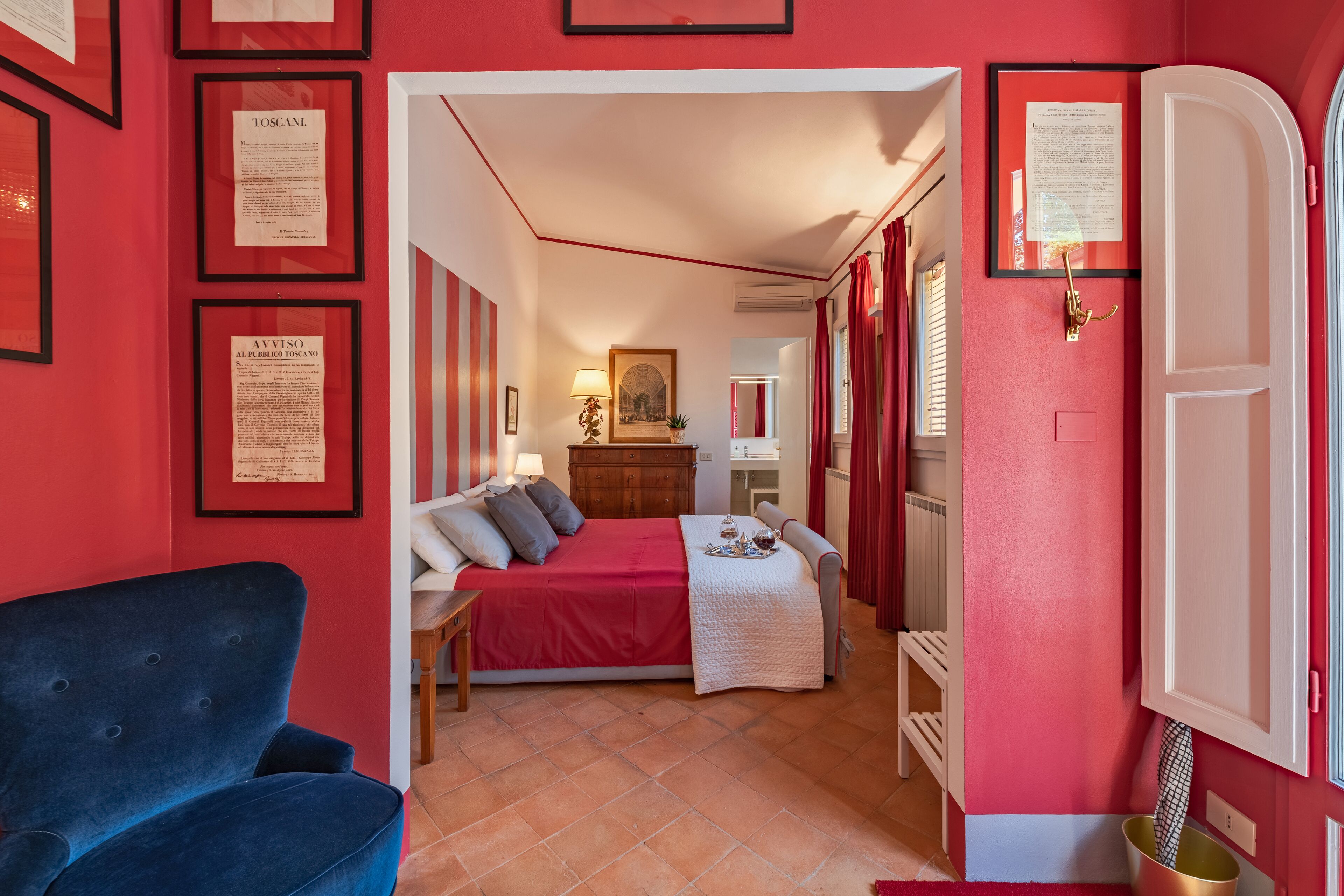 A bedroom with a bed, a blue armchair, a dresser, and framed documents on the wall.