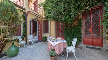 Un patio con una mesa preparada para comer, rodeado de sillas blancas, un jarrón verde grande y plantas en macetas.