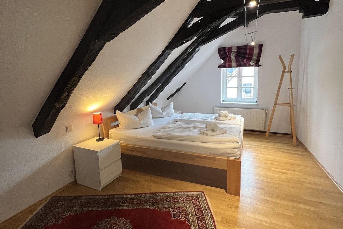 Een slaapkamer met een schuine plafond, een houten bed, een nachtkastje met een lamp, een rood gestreept tapijt en een raam met uitzicht op de omgeving.