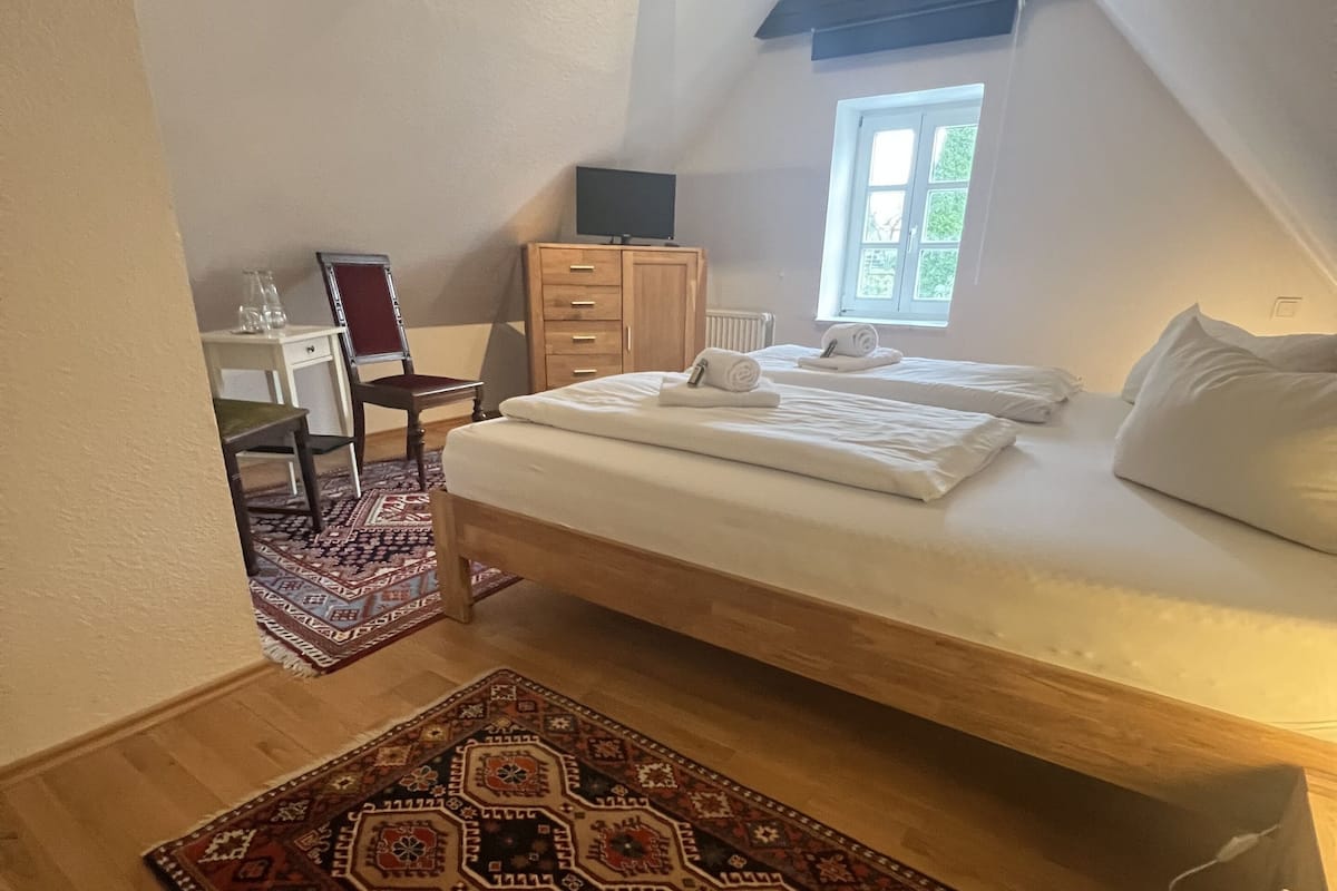 Een slaapkamer met een hoogslaper, een houten dressoir, een stoel, een klein tafeltje en een kamerkleed met patroon.