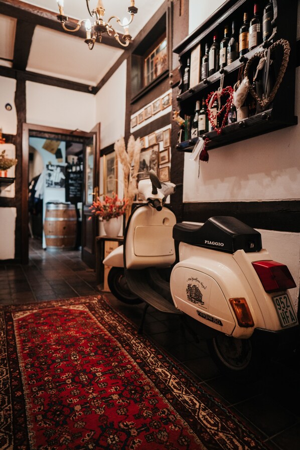 A vintage Vespa scooter parked in a hallway with a red patterned rug, wooden beams, and a wine shelf with bottles. - Altes Gasthaus Nagel (Oerlinghausen)