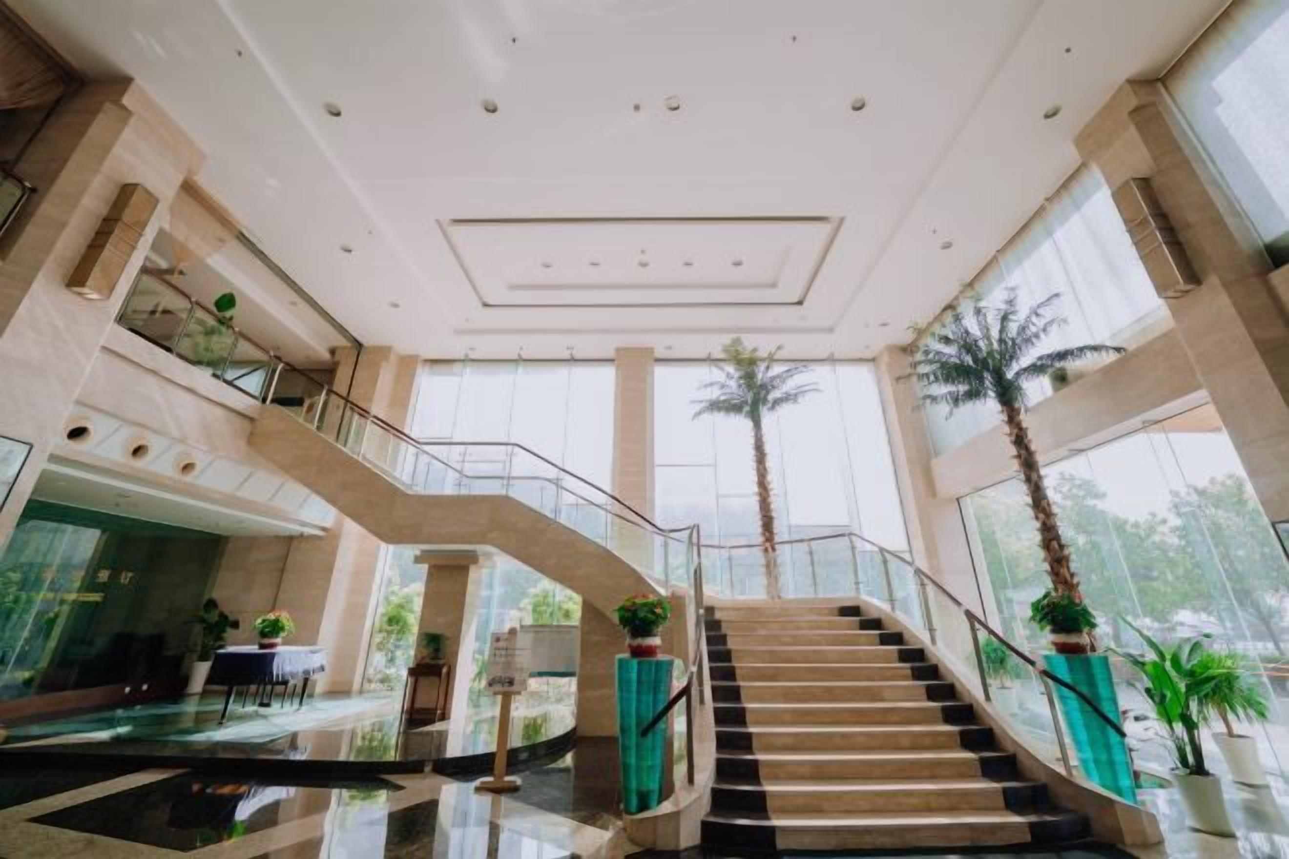 A spacious lobby with a central staircase, large windows, and indoor plants.