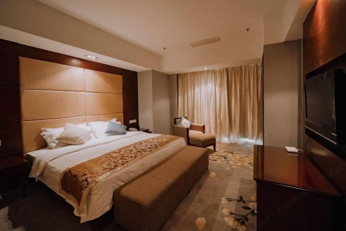 A hotel room with a large bed, a bench, a TV, a desk, and a window with curtains.
