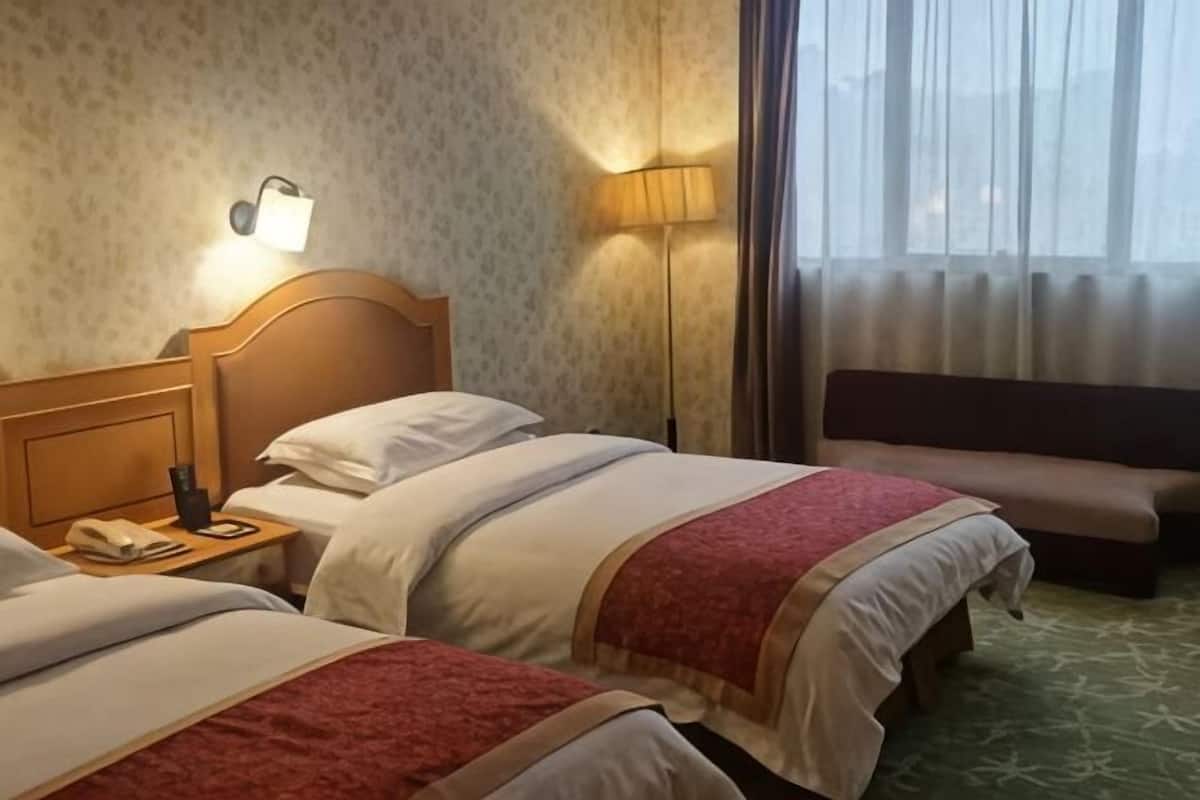 A hotel room with two beds, a nightstand, a lamp, a bench, and a view of the outdoors.