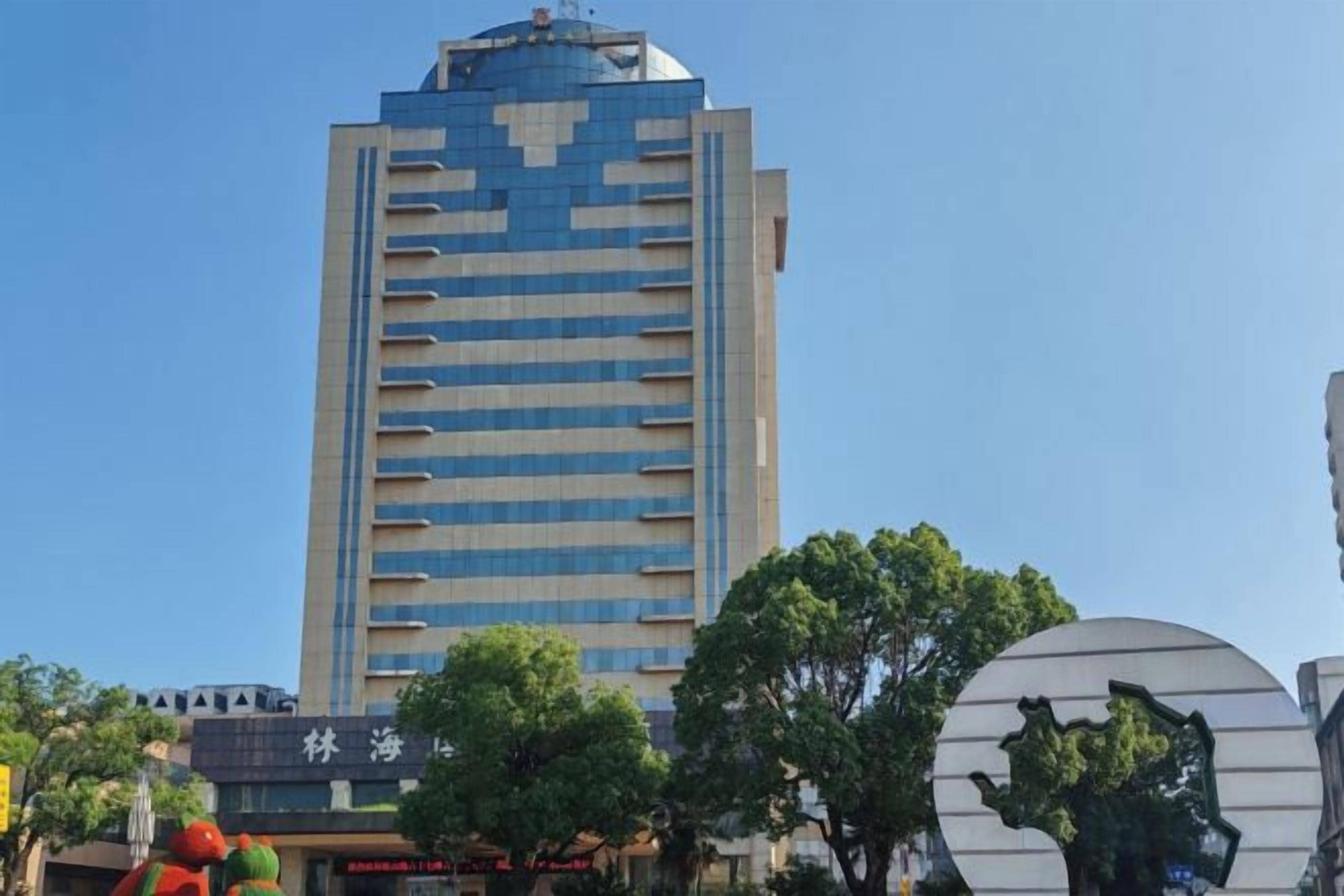 Un edificio alto con diseño moderno, rodeado de árboles y otros edificios.