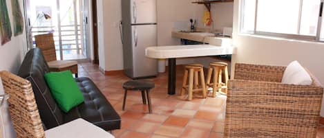 A compact living space with a sofa, a wicker chair, a small stool, a refrigerator, and a kitchen area with a sink and countertop.