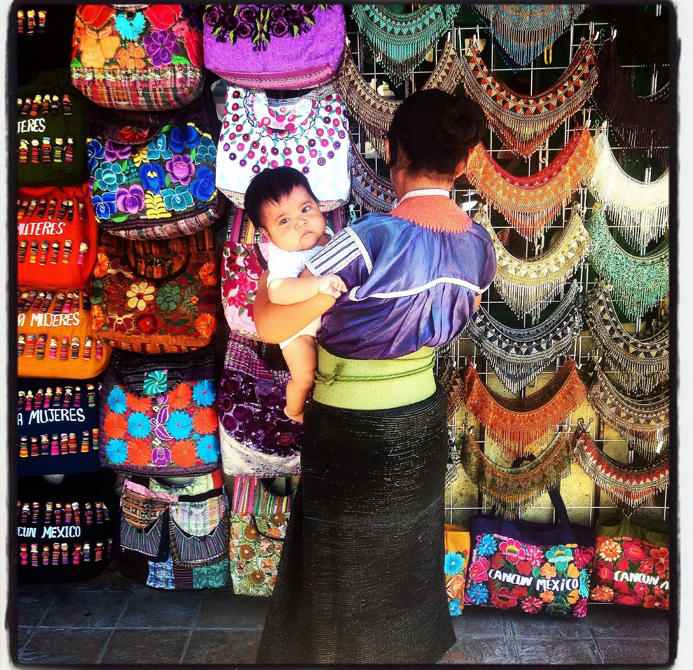 Une personne tenant un bébé devant un présentoir rempli de sacs et de textiles colorés, tissés.