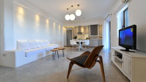 Ein modernes Wohnzimmer mit einer Couch, einem Holzstuhl, einem Esstisch mit Stühlen, einem Fernseher auf einem weißen Schrank und einer Küche im Hintergrund.