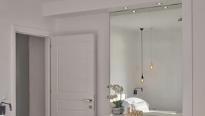 Ein Schlafzimmer mit einem Bett, einer weißen Tür, einer weißen Kommode, einem Spiegel, einer Pendelleuchte und einem Korbwäschesammler.
