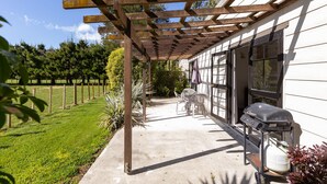 Eine überdachte Terrasse mit Grill und Stühlen, ein Betonweg und ein von Bäumen gesäumter, eingezäunter Garten.
