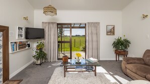 Ein Wohnzimmer mit einem Glastisch, einem braunen Sofa, einem Fernseher und Blick auf ein grünes Feld durch ein großes Fenster.