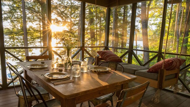 A wooden dining table set with plates, glasses, and a pitcher, surrounded by metal chairs, with a view of trees and a body of water through large windows. - Lakeside life at its best! (Enfield)