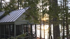 A cabin with a metal roof, large windows, and a deck overlooking a lake surrounded by trees. - Lakeside life at its best! (Enfield)