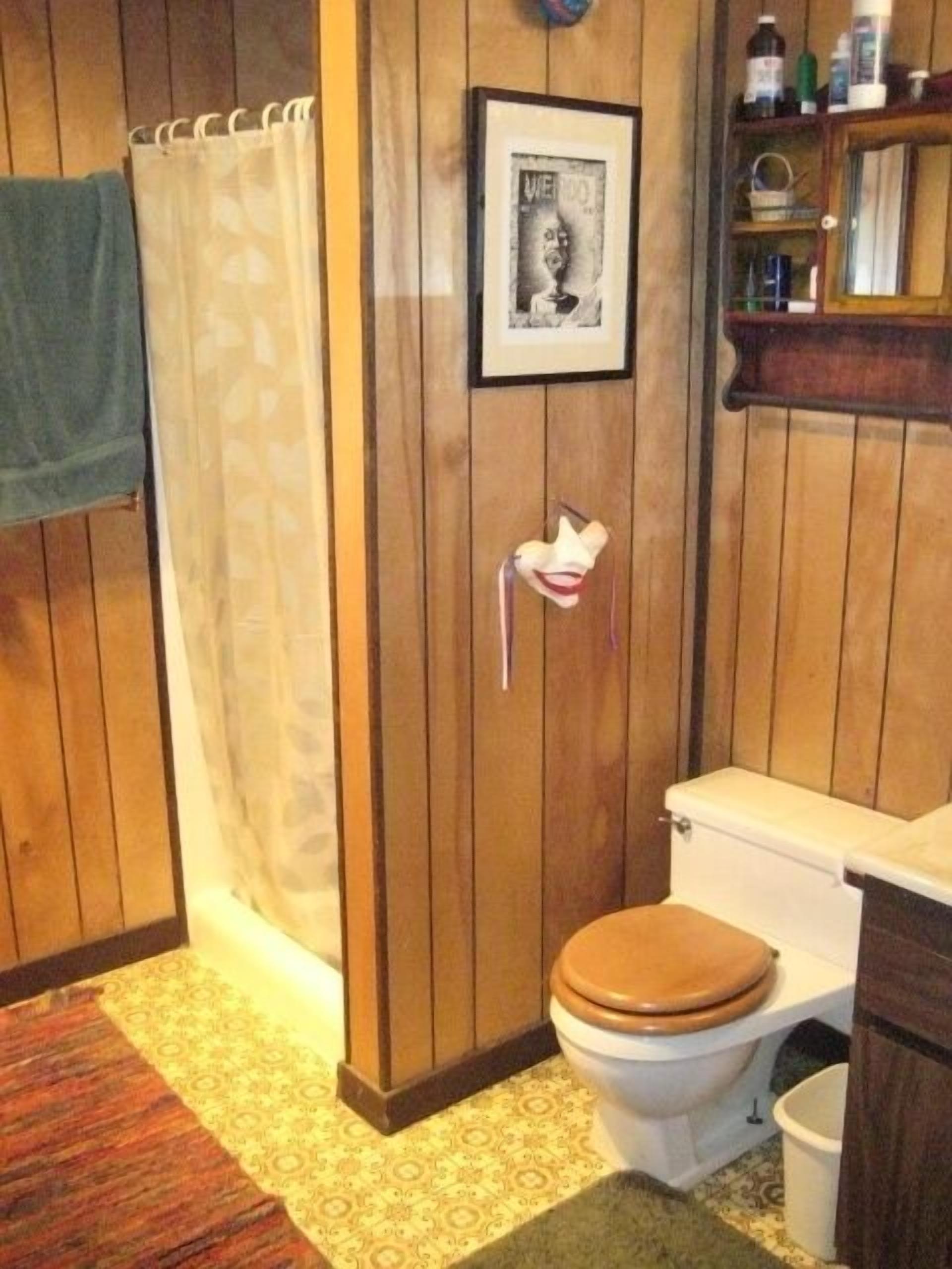 A bathroom with wooden paneling, a toilet with a wooden seat, a shower curtain, and a framed picture on the wall.