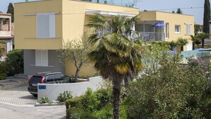 A modern two-story building with a yellow facade, a car parked in front, and a palm tree in the foreground. - Villa DANEX SUPERIOR 2 **** (Umag)