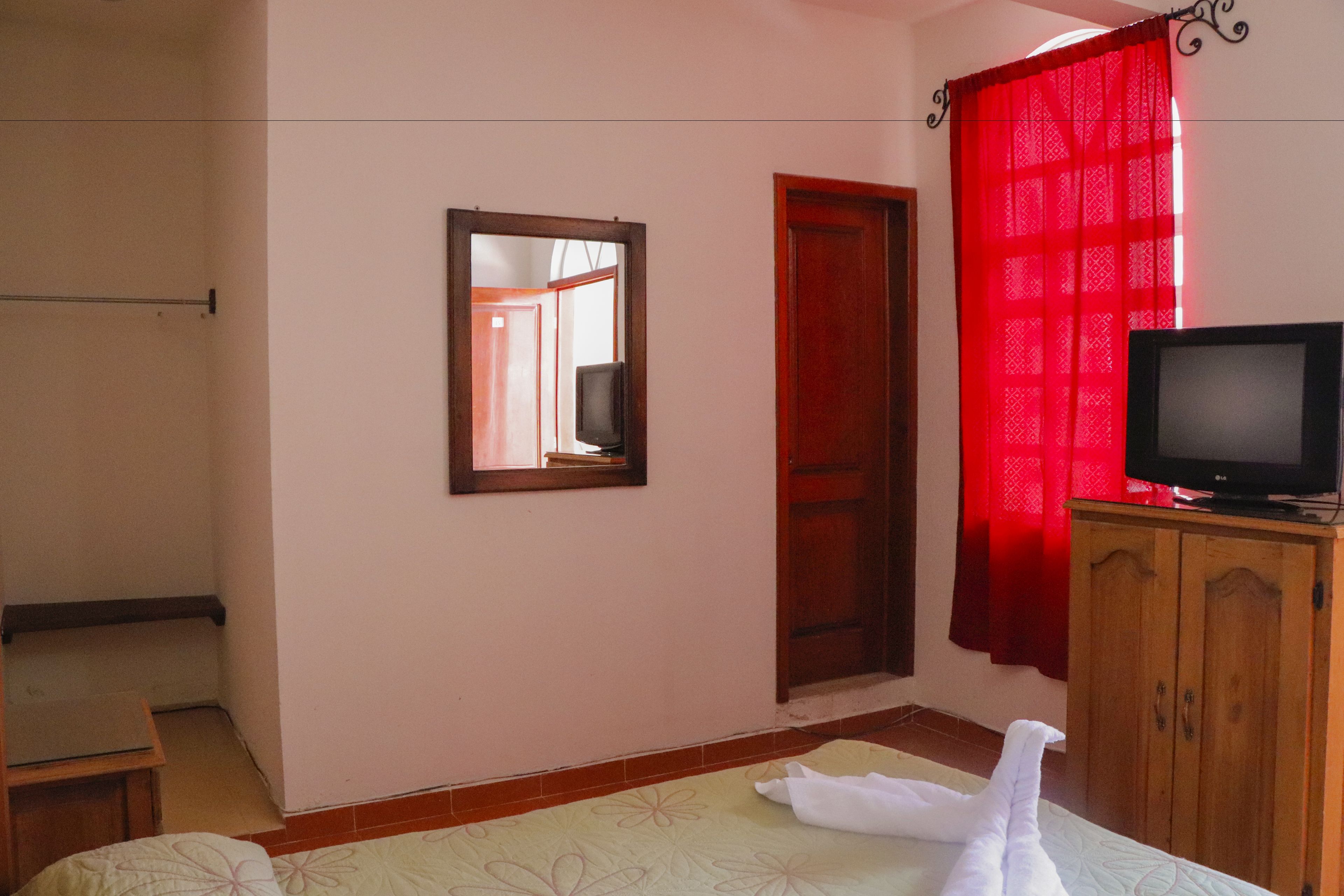 Une chambre à coucher avec un lit, un téléviseur posé sur un meuble en bois, un miroir au mur et un rideau rouge.