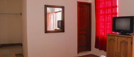 Une chambre à coucher avec un lit, un téléviseur posé sur un meuble en bois, un miroir au mur et un rideau rouge.