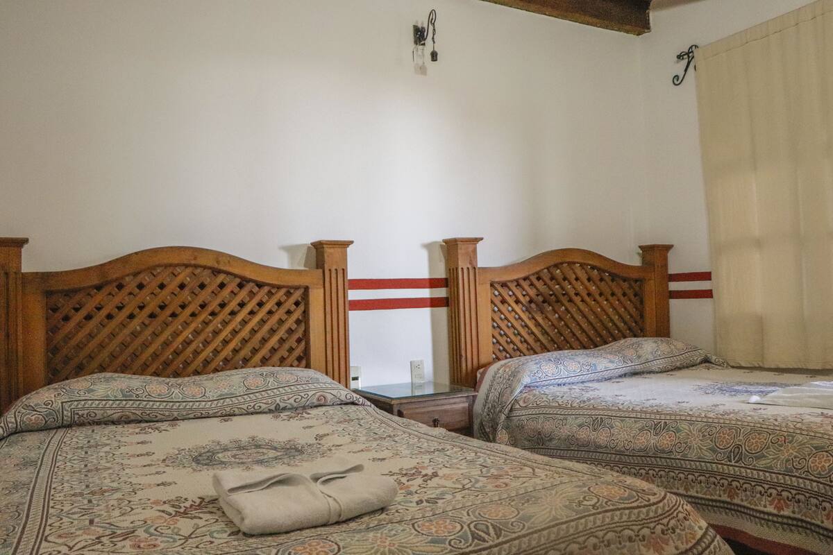 A room with two wooden beds, each with a patterned bedspread and a pillow. There is a small table between the beds, and a wall-mounted light fixture above it.