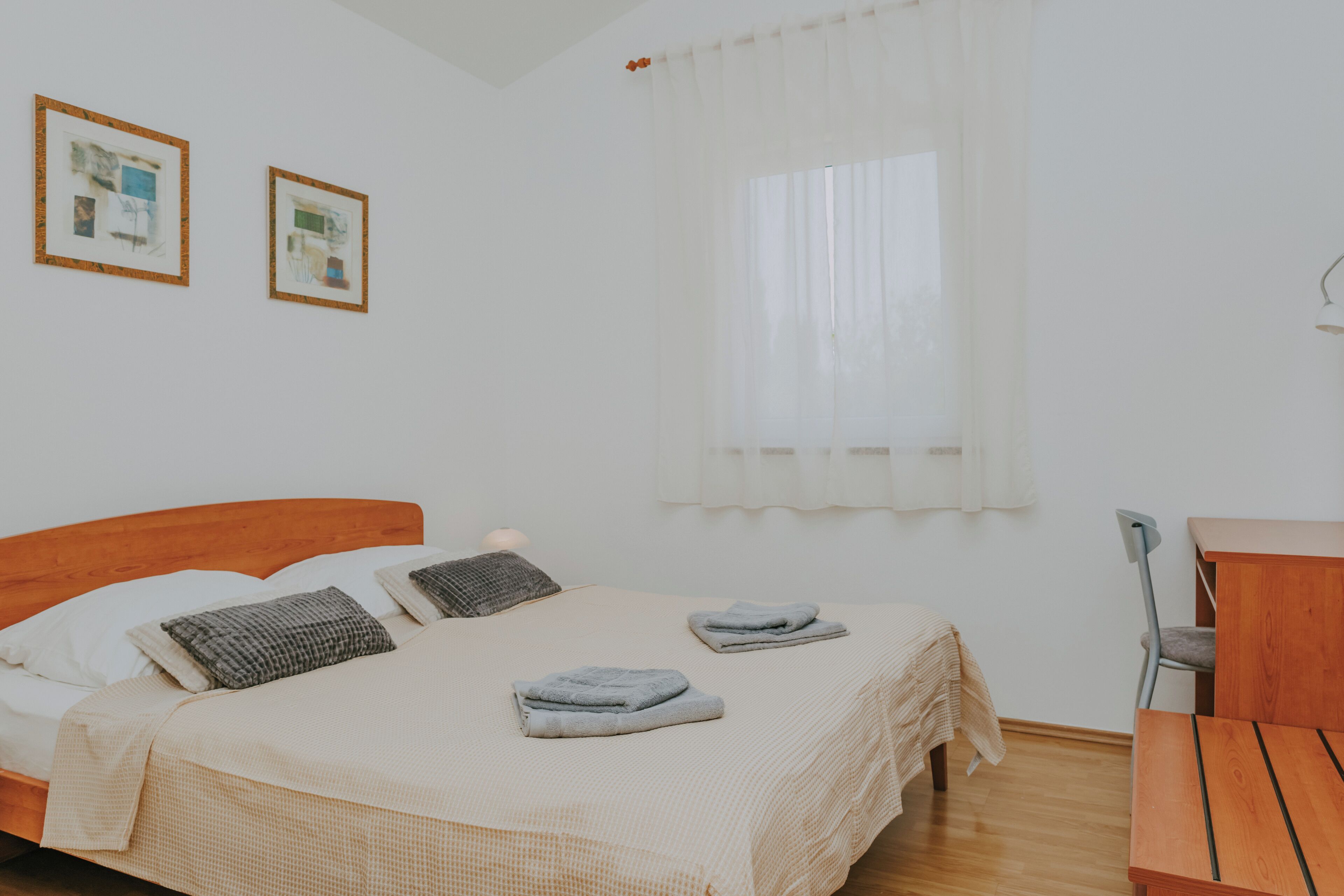 A neatly made bed with pillows and folded towels, a wooden desk, and two framed pictures on the wall.
