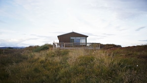 A wooden cabin with a balcony, surrounded by tall grass and shrubs. - Hestaland Riverside Cottage (Borgarbyggd)