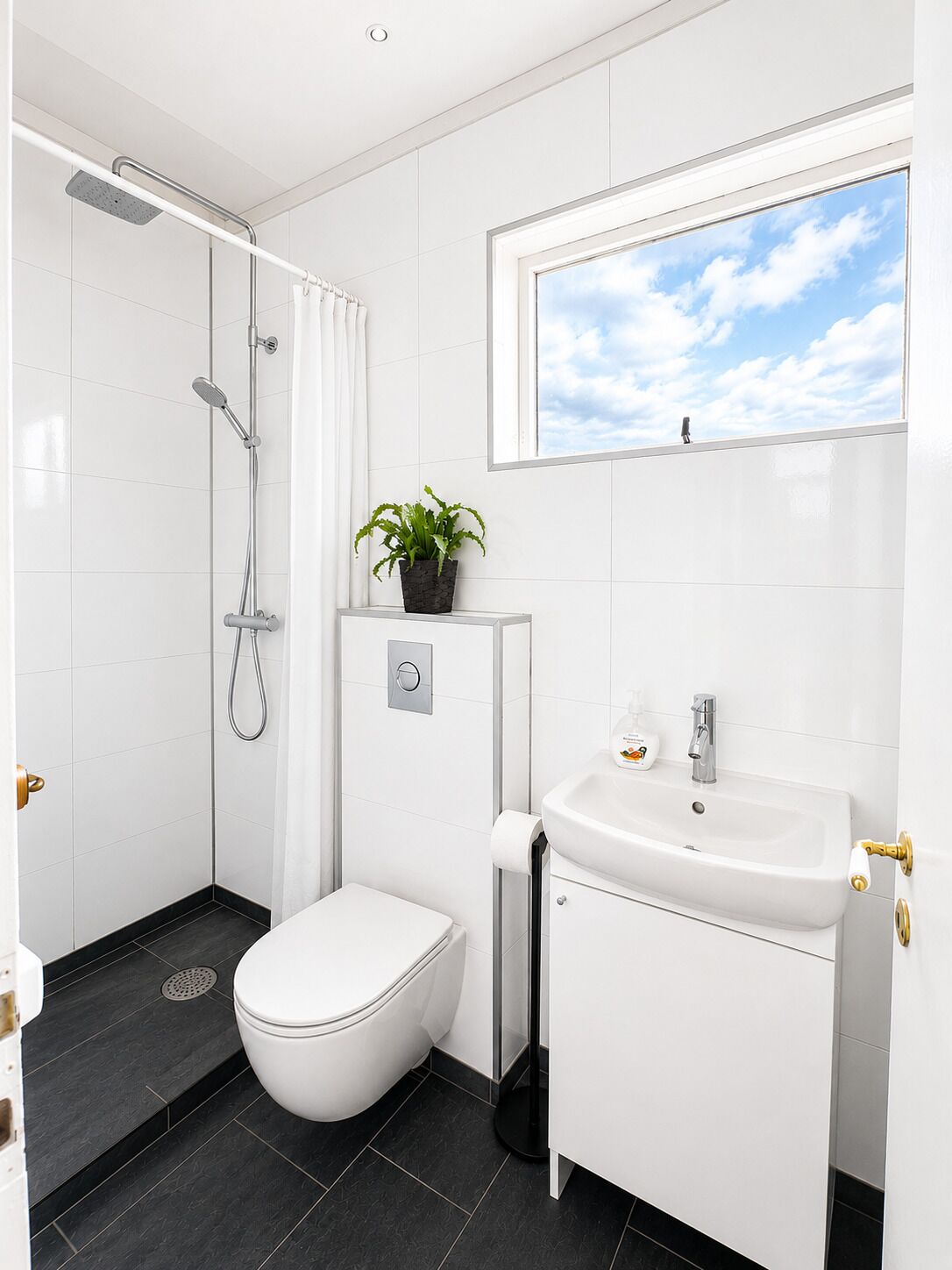 A modern bathroom with a shower, toilet, sink, and a window with a view of the sky.